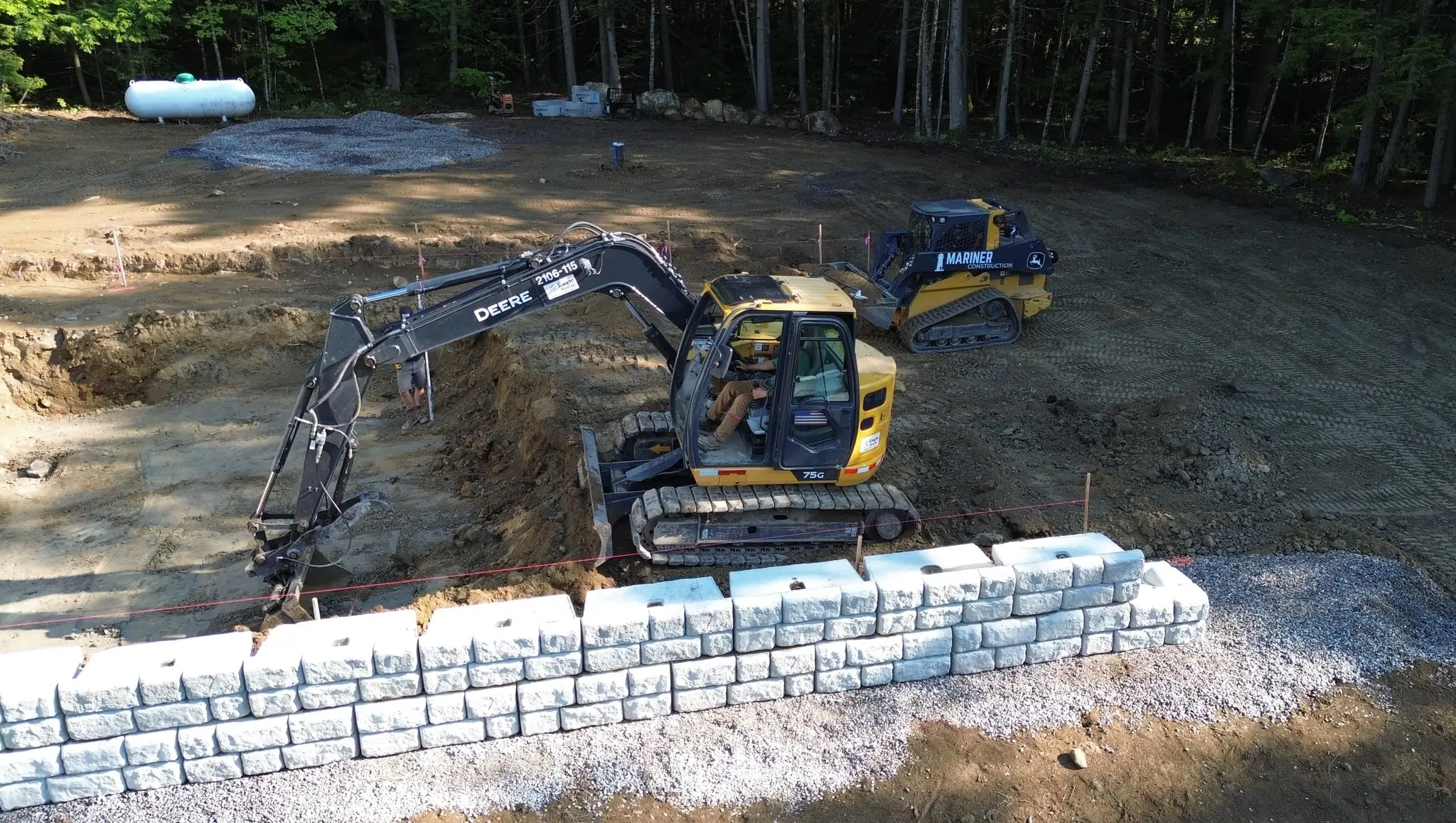 Aerial view of a Mariner Construction excavator and track loader building a block retaining wall on a Central Maine site