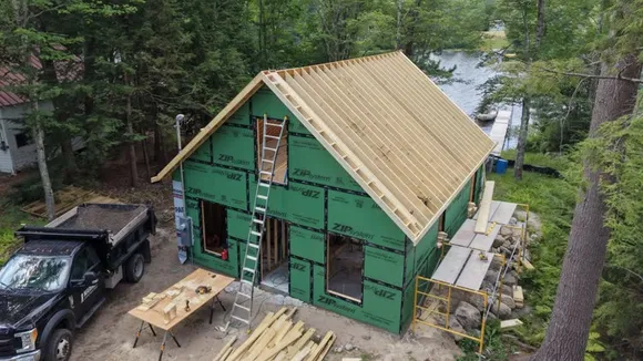 Aerial view of a new home under construction on a lakefront lot, sheathed in green ZIP System panels with roof framing underway and a Mariner Construction dump truck on site