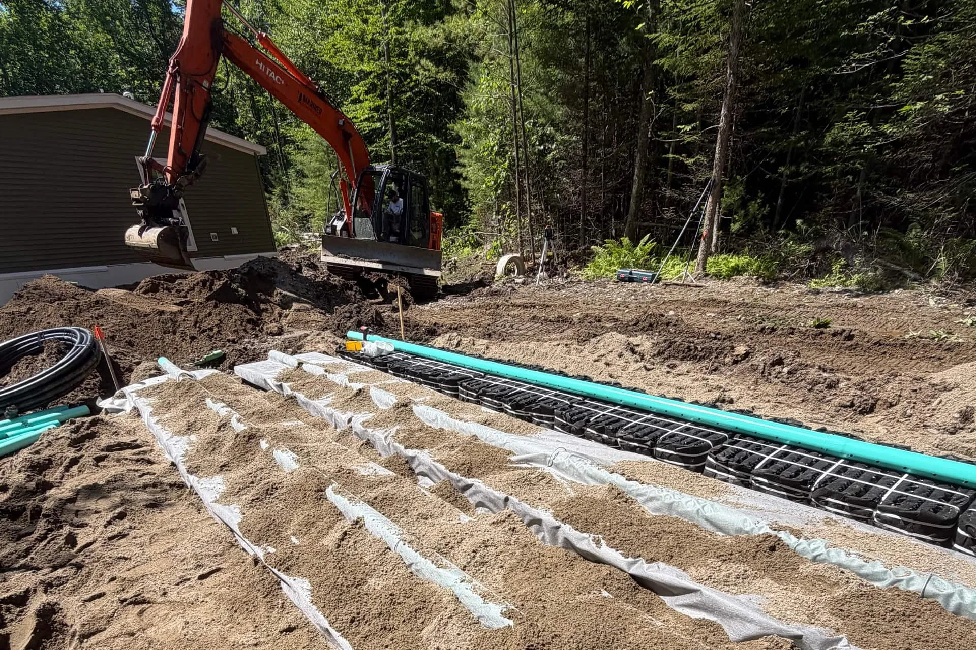 Engineered leach-field rows mid-install: sand covering geotextile fabric, a row of septic chambers and green distribution pipe still exposed, excavator working beside the house at the tree line