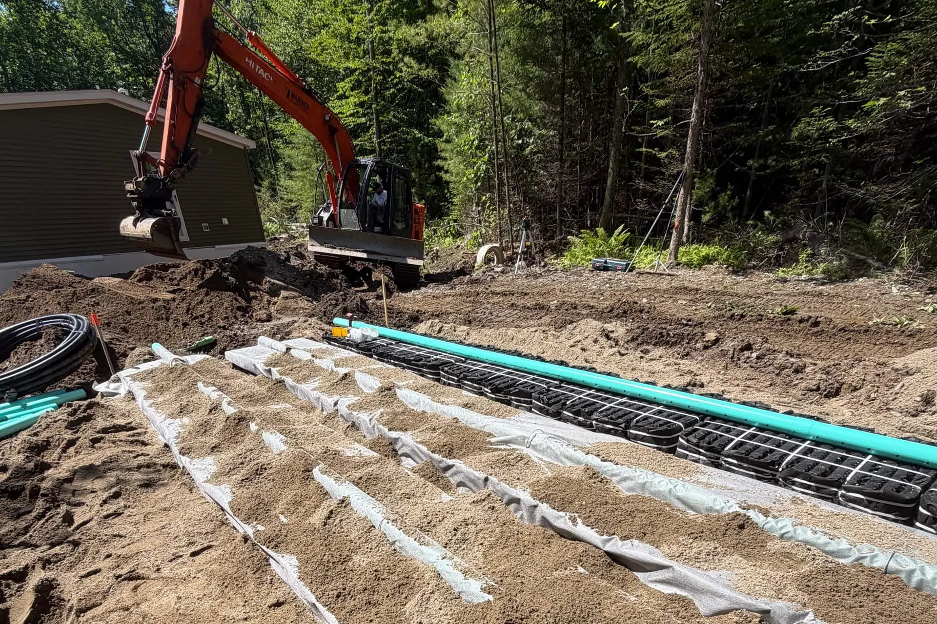 Engineered leach-field rows mid-install: sand covering geotextile fabric, a row of septic chambers and green distribution pipe still exposed, excavator working beside the house at the tree line