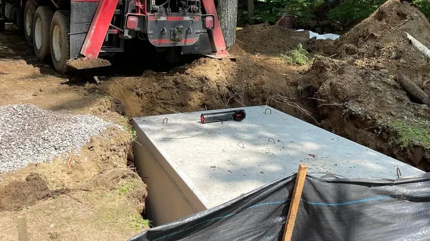 A precast concrete septic tank set in its excavation on a wooded lot, beside a boom truck