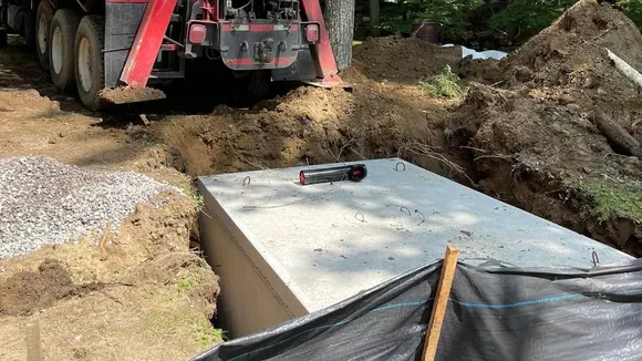 A precast concrete septic tank set in its excavation on a wooded lot, beside a boom truck