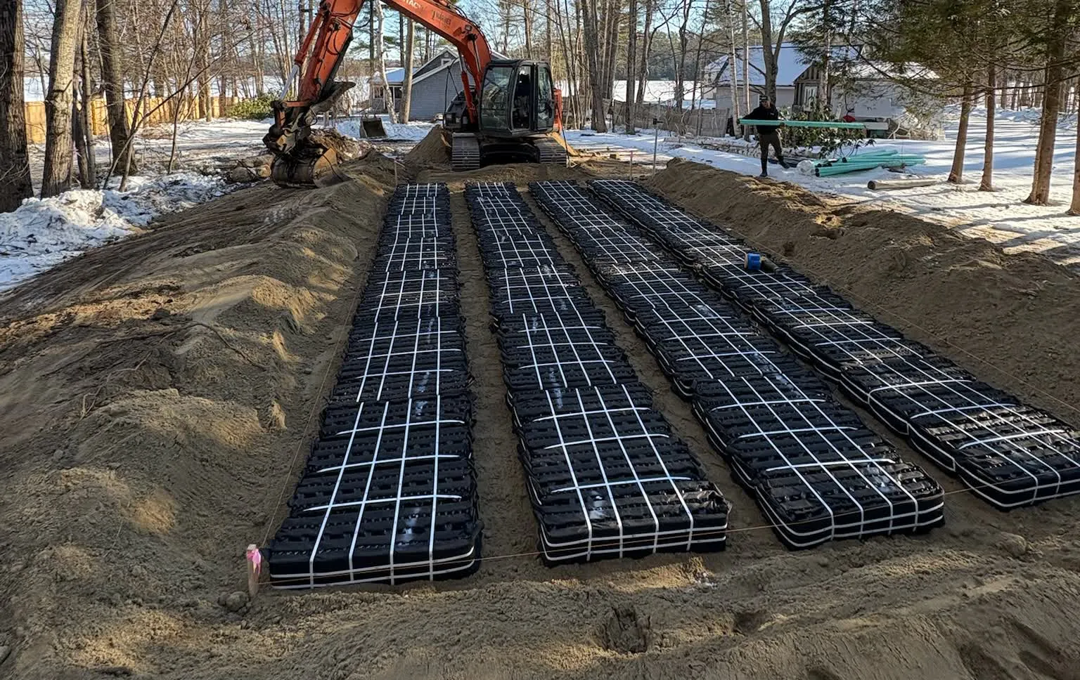 Mariner Construction crew installing rows of engineered septic leach-field modules on a sand bed during a winter install, excavator working behind them